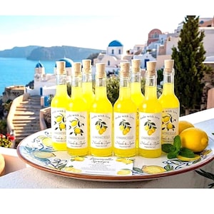 May include: Eight clear glass bottles filled with bright yellow limoncello, each topped with a cork stopper and a label reading "made with love" and "Limoncello." The bottles sit on a round ceramic platter decorated with painted lemons.