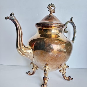Puede incluir: Una tetera de metal plateado con cuerpo abombado, pico curvo y tapa con remate decorativo. La tetera se apoya sobre tres patas ornamentadas y curvadas, e incluye un asa curva, mostrando un acabado deslustrado y reflectante.