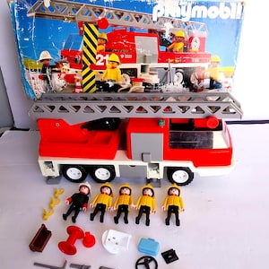 Può includere: Un camion dei pompieri giocattolo Playmobil rosso e bianco con una scala grigia estensibile, accompagnato da cinque piccole figure di vigili del fuoco in uniforme gialla e nera, insieme a vari accessori in miniatura tra cui attrezzi, un volante e un estintore.