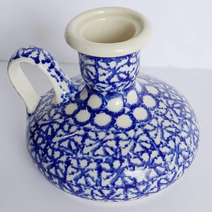 Puede incluir: Un candelabro de cerámica con base redondeada y una sola asa. La superficie presenta un patrón azul y blanco pintado con esponja que consiste en pequeñas formas florales interconectadas y motivos circulares, con un borde de color crema.