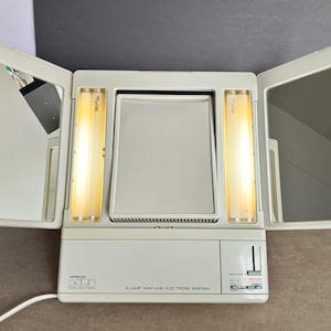 Puede incluir: Un espejo de tocador beige de tres paneles con dos alas laterales y un espejo central. La unidad cuenta con dos tubos fluorescentes verticales que flanquean el espejo central. La base tiene la etiqueta "Satin Collection" y "4 Lamp Slim Line Electronic System."