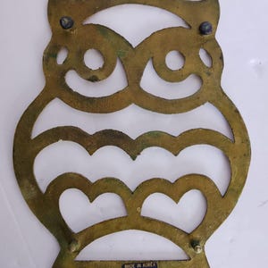 May include: A brass-colored metal trivet shaped like an owl with cut-out details for the eyes and feathers. The piece features four small feet and a blue rectangular sticker at the bottom with the text MADE IN KOREA FOR INTERPUR printed in white.
