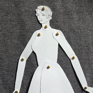 Peut inclure: Une poupée en papier blanc avec des détails dessinés à l'encre noire, dotée d'articulations mobiles reliées par de petites attaches parisiennes dorées. La silhouette comprend une tête, un torse, des bras et une jupe, découpés dans du papier.