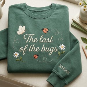 May include: A forest green crewneck sweatshirt featuring embroidered designs of a white butterfly, two ladybugs, and two white daisies. The text "The last of the bugs" is embroidered in the center, and the name "SARAH" is embroidered on the sleeve.