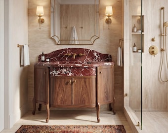 May include: A bathroom vanity with a dark red marble countertop and a wooden cabinet base, featuring a gold-framed mirror, two wall sconces, and a glass-enclosed shower with gold fixtures, all set against beige stone walls and a patterned rug.