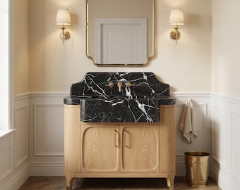 May include: A bathroom vanity featuring a black marble countertop with white veining, a light oak wood cabinet with two doors, and brass hardware. Above the vanity is a rectangular mirror with rounded corners and two brass wall sconces.