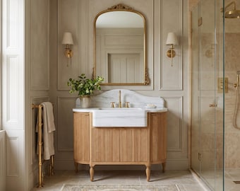 May include: A bathroom vanity with a white marble sink and countertop, featuring a light wood fluted cabinet base. Above the vanity is an arched gold-framed mirror flanked by two wall-mounted sconces. A small green plant sits on the counter.