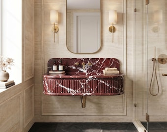May include: A wall-mounted bathroom vanity featuring a deep red marble basin with white veining and a fluted front design. The vanity includes a gold faucet, two small bottles, and a stack of folded towels, positioned below a rounded rectangular mirror.