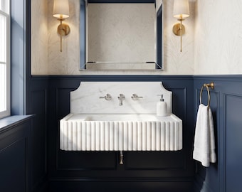 May include: A wall-mounted white marble sink with a fluted front design, featuring a matching marble backsplash, three polished chrome faucet handles, and a white soap dispenser. Two gold wall sconces with white shades flank a rectangular mirror.