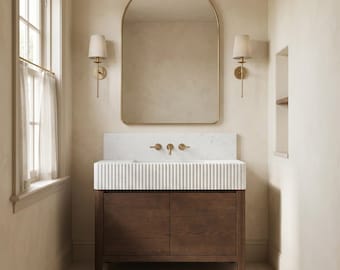 May include: A bathroom vanity featuring a dark wood cabinet with two doors, topped by a white fluted sink basin and a marble backsplash. Above, an arched mirror with a thin gold frame is flanked by two wall-mounted sconces with cream shades.