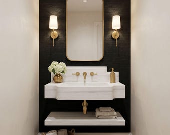May include: A wall-mounted white marble sink with a gold faucet and handles, positioned below a rectangular gold-framed mirror. Two gold wall sconces with white shades flank the mirror. A lower marble shelf holds folded towels above a woven basket.