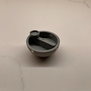 May include: A matte gray, bowl-shaped object featuring a central horizontal bar with a small circular knob attached to one end. The rim of the bowl contains several small, circular metallic inserts spaced evenly around the circumference.