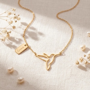 May include: A gold-toned necklace featuring a delicate hummingbird outline pendant and a small rectangular charm engraved with the name Emma. The necklace is crafted with a fine chain and is accompanied by scattered white pearls and dried flowers.