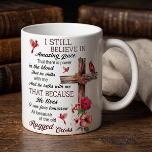 May include: A white ceramic mug featuring a printed design of a wooden cross adorned with pink hibiscus flowers and red cardinal birds. The text reads: I STILL BELIEVE IN Amazing grace That there is power in the blood That he shdks with me And he twlks with me THAT BECAUSE He lives I can face tomorrow All because of the old Rugged Cross.