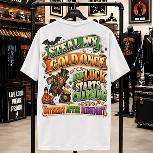 Pode incluir: Uma t-shirt branca com uma impressão gráfica de um duende com um pote de ouro, uma abóbora e um morcego. O texto diz: STEAL MY GOLD ONCE AND LUCK STARTS CHARGING INTEREST AFTER MIDNIGHT. O design inclui detalhes em verde, laranja e dourado.