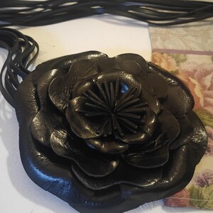 May include: A black leather flower necklace featuring multiple layered petals and a central cluster of thin, upright strips. The piece is attached to several thin black leather cords that extend from the back of the floral centerpiece.