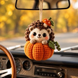 May include: A crocheted hedgehog ornament hanging from a string, featuring a cream-colored face, dark brown textured back, and pink cheeks. The hedgehog sits inside a bright orange crocheted pumpkin with a small orange mushroom and green leaves.