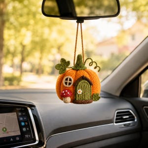 May include: A crocheted pumpkin-shaped hanging ornament featuring a green stem, a small arched door, a circular window, and a red-and-white spotted mushroom. The orange pumpkin is suspended by a brown cord from a vehicle rearview mirror.