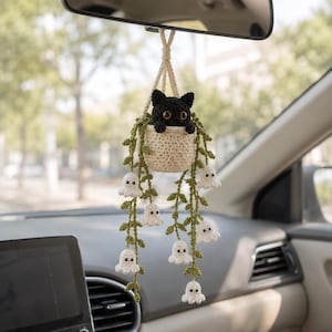 May include: A crocheted car accessory featuring a black cat with large eyes sitting in a cream-colored pot. Green vines hang down from the pot, decorated with small white crocheted ghost figures. The entire piece is suspended by a cream-colored cord.