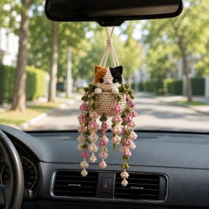 May include: A crocheted calico cat ornament sitting in a small beige basket, suspended by a cord. The cat has black, orange, and white fur. Cascading from the basket are green vines with small, delicate pink and white crocheted flower clusters.