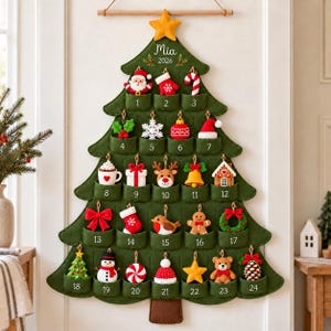 May include: A green felt Christmas tree wall hanging advent calendar with 24 numbered pockets. Each pocket contains a small felt ornament including a Santa, snowflake, reindeer, and gingerbread man. The top features a yellow star and the text Mia 2026.