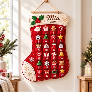 May include: A red felt Christmas stocking advent calendar featuring the name Mia and the year 2026 embroidered at the top. It has twenty-four numbered pockets, each containing a small festive ornament like a Santa, tree, or star.