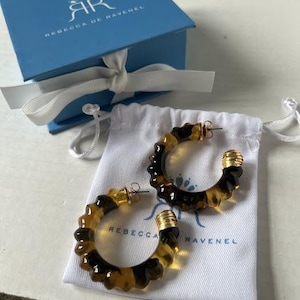 May include: A pair of hoop earrings featuring a translucent brown tortoiseshell pattern with a segmented, bamboo-like texture. Each earring has a gold-tone metal post and a matching gold-tone metal cap at the end, resting on a white fabric pouch.