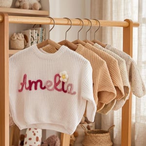 May include: A white long-sleeve knit sweater featuring the name Amelia embroidered in pink and red yarn with a small white flower accent, hanging on a wooden hanger alongside several other neutral-toned knit sweaters on a wooden clothing rack.