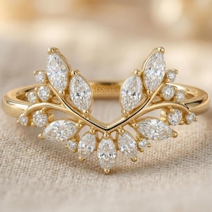 Può includere: Un anello in oro 18 carati con una fascia a forma di V decorata con un gruppo di diamanti bianchi taglio marquise e rotondo. La fascia in metallo presenta l'incisione 18K 750 sulla superficie interna, mostrando una disposizione simmetrica.
