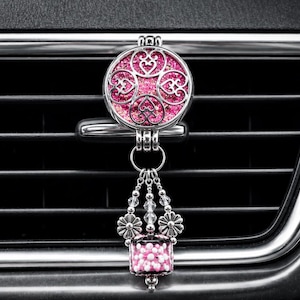 May include: A silver-toned car air freshener vent clip featuring a circular locket with a glittery pink heart-patterned inlay. A dangling charm hangs from the locket, consisting of small silver flower beads and a rectangular glass bead filled with pink spheres.