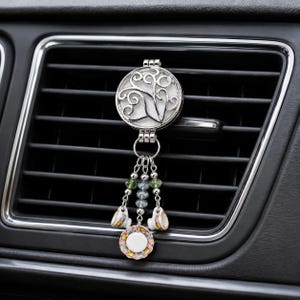 May include: A silver-toned car air freshener vent clip featuring a circular locket with a white inlay and a swirling metal design. Three dangling chains hang from the locket, adorned with small green and blue beads, two miniature teacups, and a saucer.