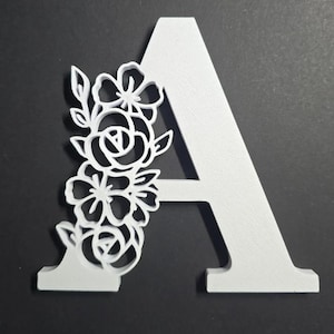 May include: A white, three-dimensional letter A featuring a decorative floral cutout design attached to the left side. The floral pattern consists of stylized flowers and leaves with open spaces, creating a layered effect on the solid letter.