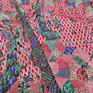 Puede incluir: Una tela roja vibrante que presenta un patrón batik detallado con aves estilizadas, motivos florales y formas geométricas en tonos azul, verde, negro y blanco. El diseño intrincado cubre toda la superficie con capas superpuestas.
