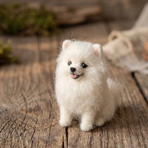 May include: A small, white, needle-felted Pomeranian dog figurine with fluffy, textured wool fibres, dark bead eyes, a black nose, and a tiny open mouth showing a pink tongue, standing in an alert pose with upright ears and a thick, soft coat.