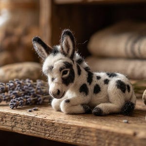 May include: A small needle-felted donkey figurine with white fur and black spots. The donkey is in a lying position with its ears upright, featuring dark markings around its eyes and ears, and a small black tail, crafted with a soft, textured appearance.