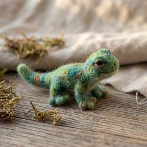 May include: A needle-felted lizard figurine crafted from wool fibers in shades of lime green, forest green, and bright blue. The small creature features a textured, fuzzy surface, a long tail, four distinct legs, and dark, round bead eyes.