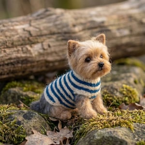 May include: A needle-felted Yorkshire Terrier figurine with tan and gray fur, wearing a cream-colored sweater featuring horizontal navy blue stripes. The small dog sculpture has dark eyes, a black nose, and pointed ears, sitting in a realistic pose.