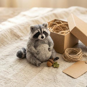 May include: A needle-felted raccoon figurine with gray and white fur, black eye patches, and a striped tail. The raccoon sits next to a small brown acorn with a green leaf, a cardboard gift box filled with straw, a coil of twine, and a blank tag.