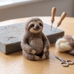 May include: A needle-felted sloth figurine made of brown and cream wool fibers. The sloth has dark eyes, a small nose, and a subtle smile. Beside it are two felting needles with wooden handles, a grey foam block, wool roving, and small scissors.