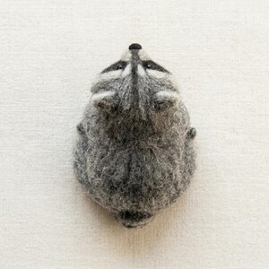 May include: A needle-felted raccoon figurine crafted from textured gray and white wool fibers. The raccoon is depicted from a top-down perspective, showcasing its pointed ears, dark eye patches, and a small black nose on a rounded head.