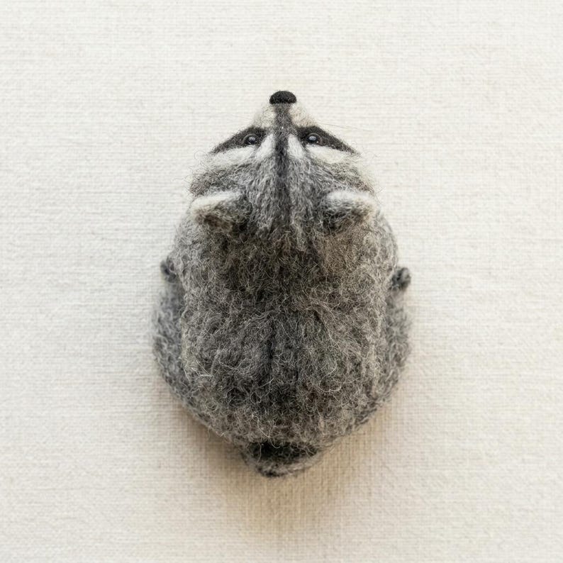 May include: A needle-felted raccoon figurine crafted from textured gray and white wool fibers. The raccoon is depicted from a top-down perspective, showcasing its pointed ears, dark eye patches, and a small black nose on a rounded head.