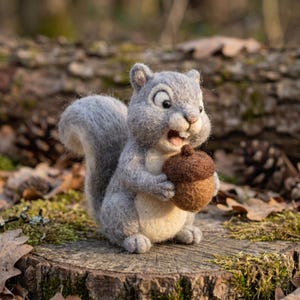 May include: A needle-felted gray squirrel figurine with a cream-colored belly, black eyes, and a small open mouth. The squirrel has a bushy tail and is holding a textured brown acorn with a dark brown cap in its front paws.