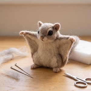 May include: A needle-felted flying squirrel figurine crafted from light brown and cream-colored wool fibers. The small creature features dark, round eyes, pointed ears, and extended patagium membranes, positioned with two metal needles and scissors.