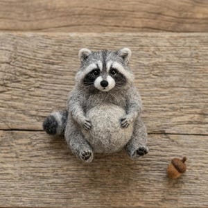 May include: A needle-felted raccoon figurine with gray and white fur, a black eye mask, and a striped tail, sitting upright with a round belly. A small brown felted acorn is positioned to the lower right of the raccoon figure.