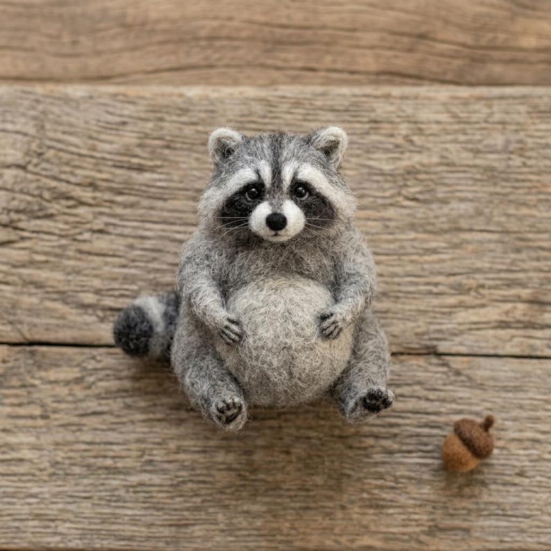 May include: A needle-felted raccoon figurine with gray and white fur, a black eye mask, and a striped tail, sitting upright with a round belly. A small brown felted acorn is positioned to the lower right of the raccoon figure.