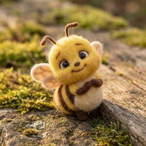 Pode incluir: Uma figura de abelha em lã feltrada com um corpo redondo às riscas amarelas e castanhas, pequenas asas claras, duas antenas e grandes olhos escuros. O objeto artesanal de lã texturizada tem uma expressão sorridente e está de pé.