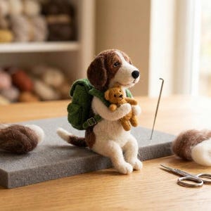 May include: A needle-felted beagle figurine with brown and white fur, wearing a green backpack and holding a small brown teddy bear. The dog is crafted from wool fibers and features a black nose, positioned on a gray foam mat with a needle.