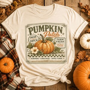 Puede incluir: Una camiseta de color crema con un estampado gráfico de una calabaza naranja grande con hojas verdes y una flor blanca. El texto dice PUMPKIN Patch, FRESH FROM THE PATCH, FARM FRESH LOCALLY GROWN y PUMPKINS CORN MAZE APPLE CIDER.