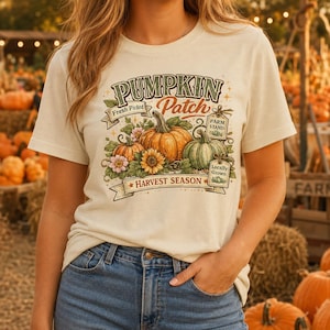 Puede incluir: Una camiseta color crema con un estampado gráfico de calabazas, girasoles y hojas. El texto dice PUMPKIN PATCH, Fresh Picked, PARM STAND, LOCALLY GROWN y HARVEST SEASON. El diseño incluye calabazas de color naranja y verde con flores.
