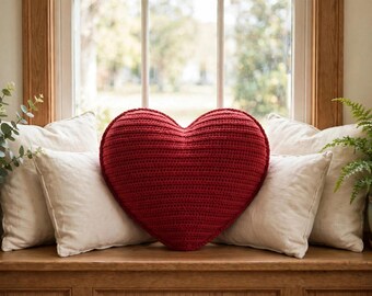 May include: A large, heart-shaped pillow with a deep red, textured crochet knit surface. The pillow is positioned centrally, flanked by four plain, off-white square throw pillows on a wooden ledge in front of a window.